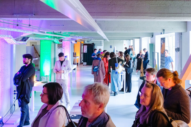 Das Bild zeigt einen hellen, weißen Raum mir ein paar farblich beleuchteten Flächen, in dem sich ca 20 Personen eine Ausstellung ansehen.