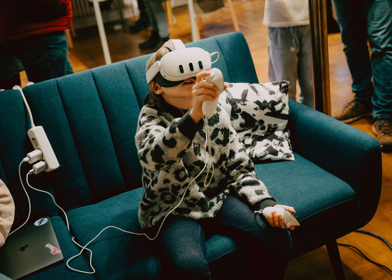 Das Bild zeigt ein Kind auf einem dunkelblauen Sofa, welches eine VR Brille trägt und Joysticks in den Händen hält.