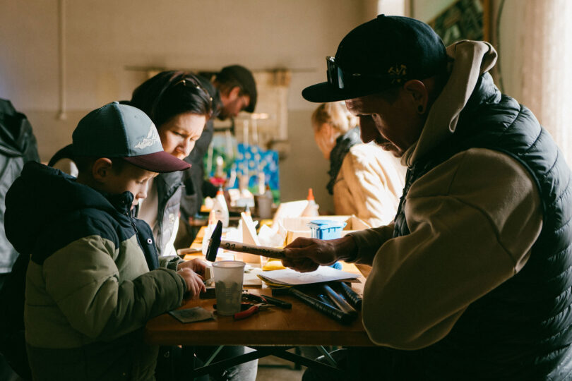 Das Bild zeigt mehrere Personen (vermutlich Eltern mit Kind), die an etwas mit Holz arbeiten. Der Papa hat einen Hammer in der Hand.