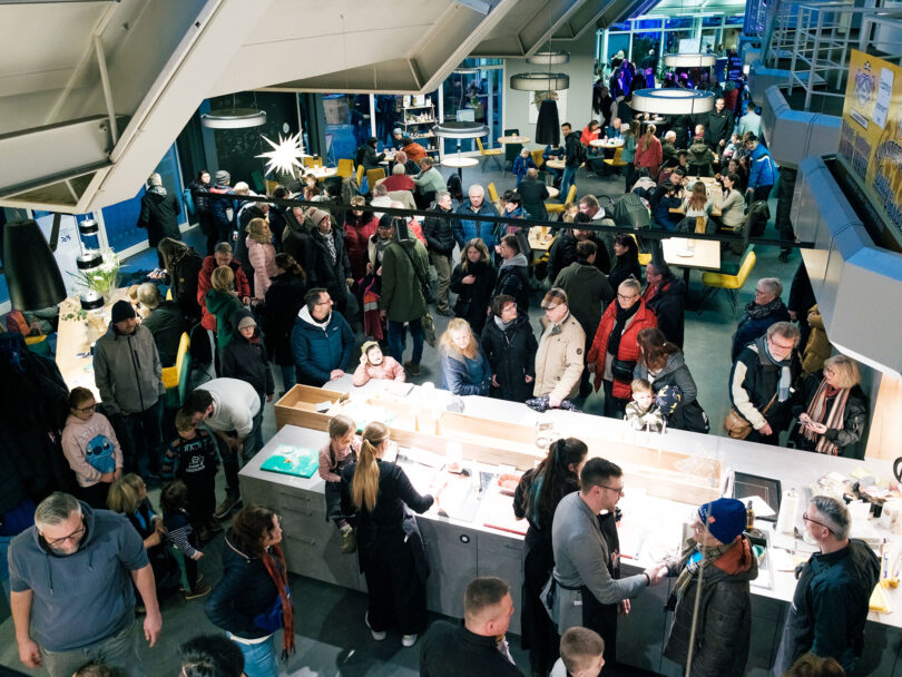 Das Bild zeigt viele Personen in einer größeren Halle stehend und sitzend von der Empore fotografiert.