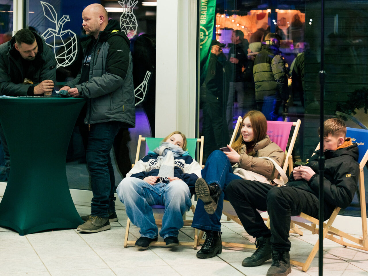 Das Bild zeigt fünf Personen. Zwei männliche Personen stehen an einem Tisch und drei jugendliche Personen liegen in Liegestühlen.