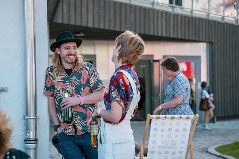 Das Bild zeigt mehrere Personen im Hof der Stadtwirtschaft in Cehmnitz. Im Vordergrund ist eine männliche (Lange helle Haare, schwarzer Hut, Getränk in der hand) und eine weibliche (buntes Hemd, Latzhose, kurze braune Haare) Person, die freudig miteinander sprechen.