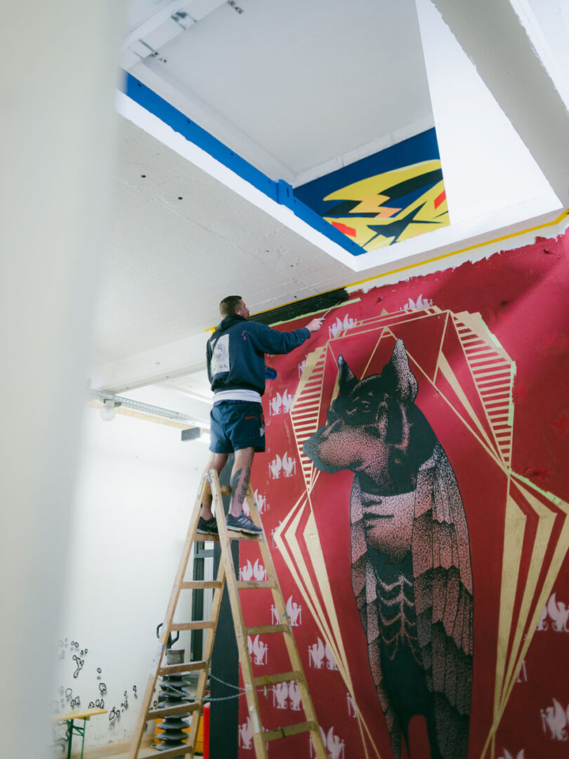 Das Bild zeigt einen Künstler auf einer Leiter, der ein großs Wandbild malt.
