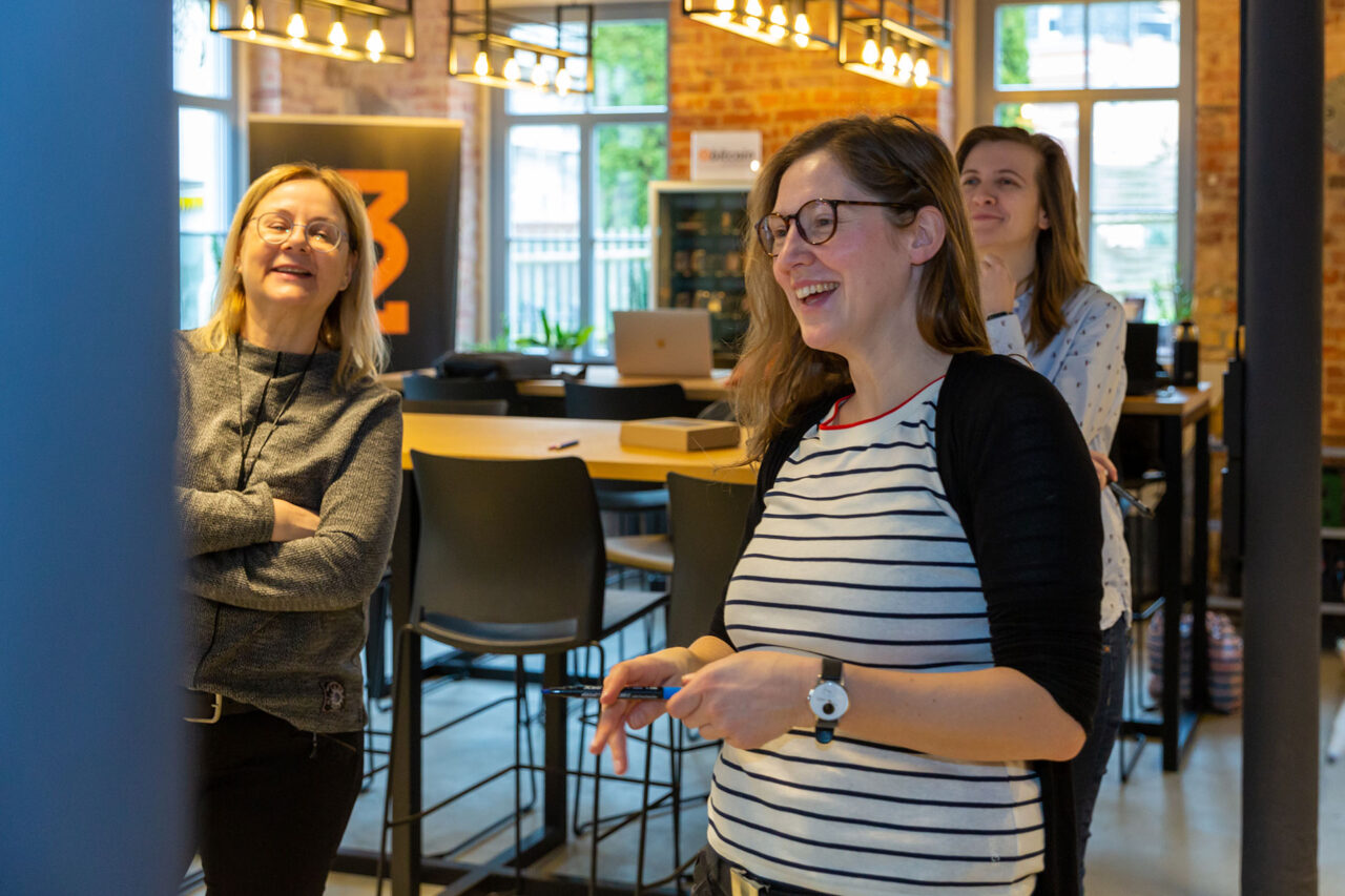 Das Bild zeigt drei weibliche, lächelnde Persone, die auf etwas schauen. Die Umgebung ist ein moderner Gastro- oder Meeting-Raum mit Hochstühlen an langen Holztischen.
